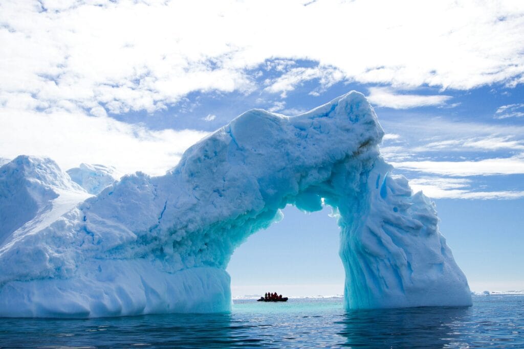 Luxury expedition ship approaching Antarctic iceberg at sunrise with kayakers exploring crystal-clear blue waters in foreground
