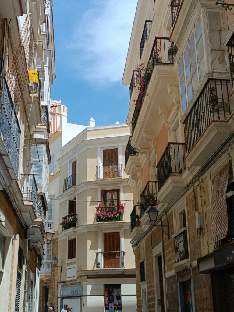 Narrow cobblestone street in Cádiz with traditional whitewashed buildings, ornate balconies, and authentic Andalusian architecture, minimal tourists visible
