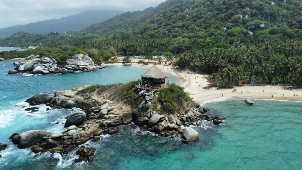 Tayrona National Park showing jungle-covered mountains meeting pristine Caribbean beach, palm trees, turquoise water, hiking trail visible through forest