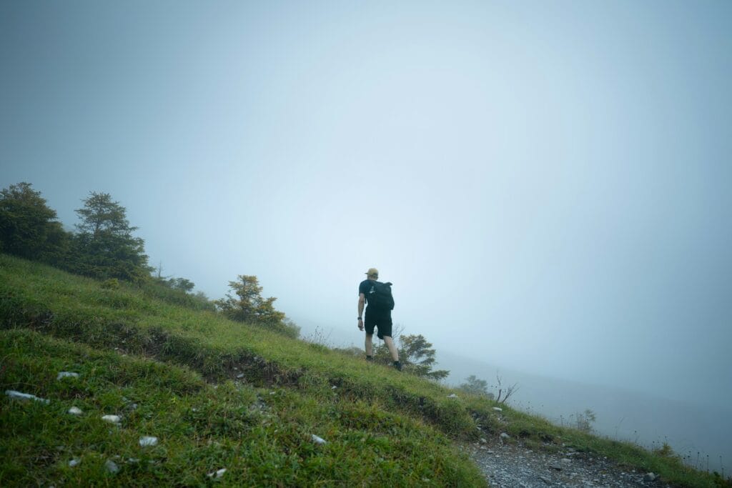 Hiker with backpack ascending foggy Alpine meadow trail, Grindelwald summer hiking, misty mountain landscape