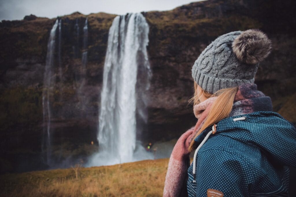 Solo female traveler with backpack and hat standing confidently at Seljalandsfoss waterfall in Iceland, looking at nature views