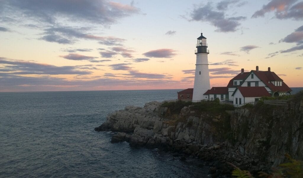 Classic Maine lighthouse on rocky peninsula at golden hour with crashing waves, pine trees, and sailboat visible in distance