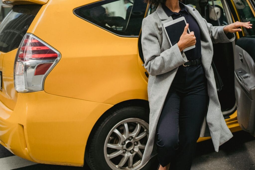 Solo female traveler with dark curly hair wearing gray blazer and holding tablet standing next to yellow taxi cab, checking rideshare details before entering