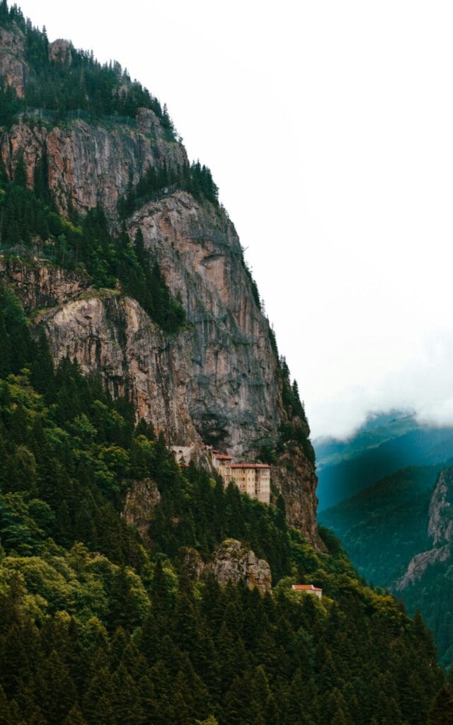 Sumela Monastery perched dramatically on clifftop surrounded by lush green mountains, Black Sea visible in distance, atmospheric mist
