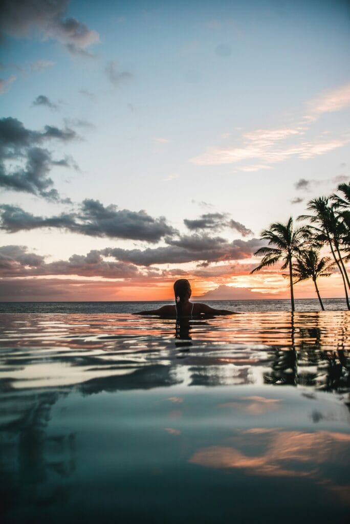 Woman swimming in luxurious infinity pool overlooking ocean at tropical resort with palm trees visible
