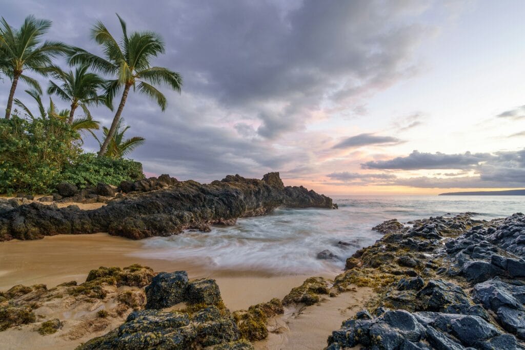 Long exposure photograph of waves crashing against a rocky volcanic shoreline in Hawaii at twilight.