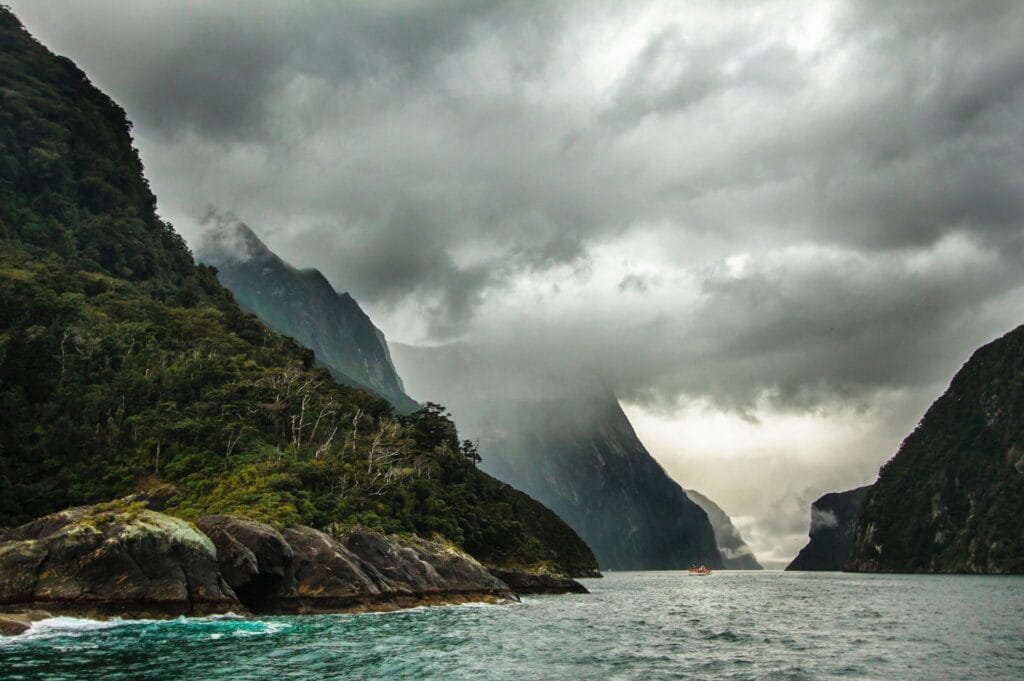 A dramatic, cloud-shrouded mountain peak rising steeply from the dark waters of Milford Sound in New Zealand.