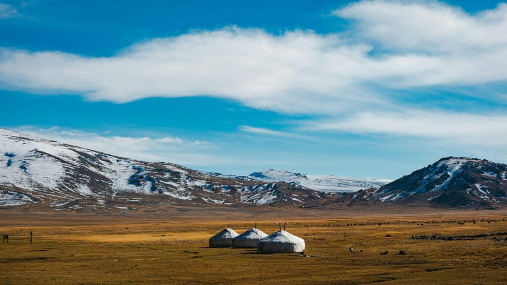 Three white traditional yurts (gers) sitting on a vast open steppe before distant snowy mountains in Mongolia