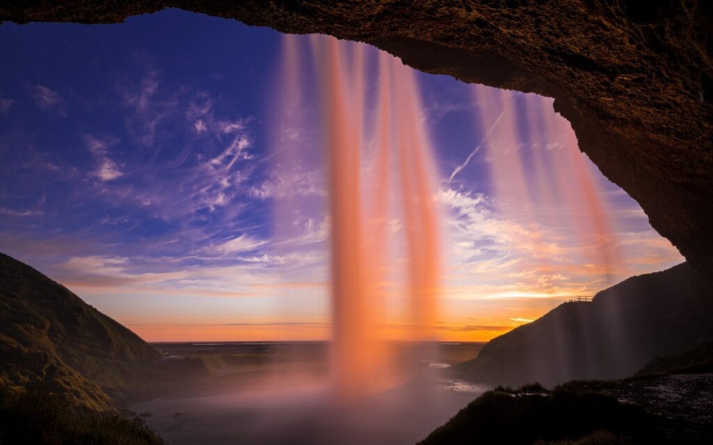 View from behind the Seljalandsfoss waterfall in Iceland during a golden sunset with a purple-streaked sky.