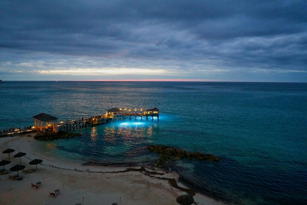 A lighted pier with cottages over clear turquoise water in Nassau, Bahamas, at dusk.