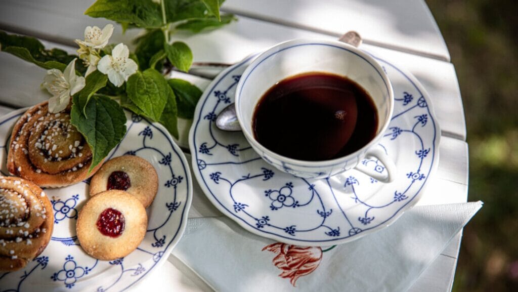 A delicate blue and white porcelain cup filled with black coffee sitting on a white garden table next to a plate of fresh kanelbullar cinnamon buns and jam cookies