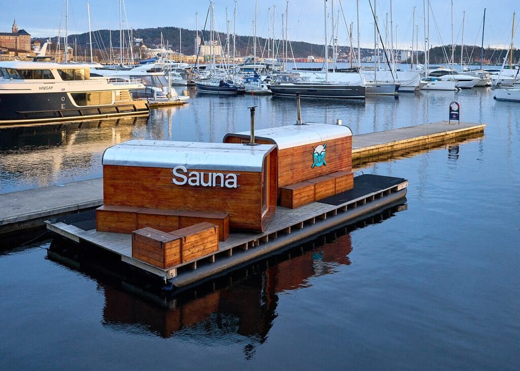 A wood-paneled KOK floating sauna structure docked at a harbor pier along the Oslo Fjord with yachts and sailboats resting in the background.