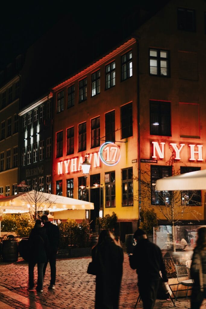 Evening view of the historic Nyhavn waterfront district during a Copenhagen hygge travel excursion, featuring illuminated neon signs for Nyhavn 17 and cozy outdoor terrace dining spaces.