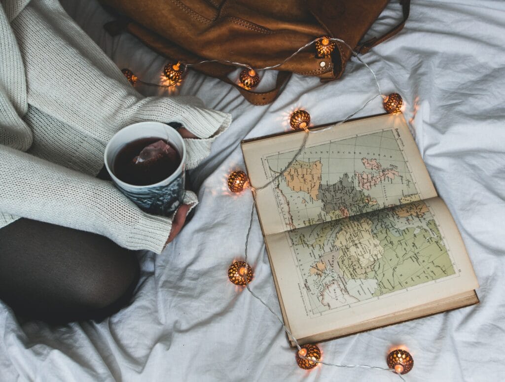 A person in a cream-colored knit sweater sitting on a bed holding a warm cup of tea next to an open vintage book map illuminated by warm glowing string fairy lights.