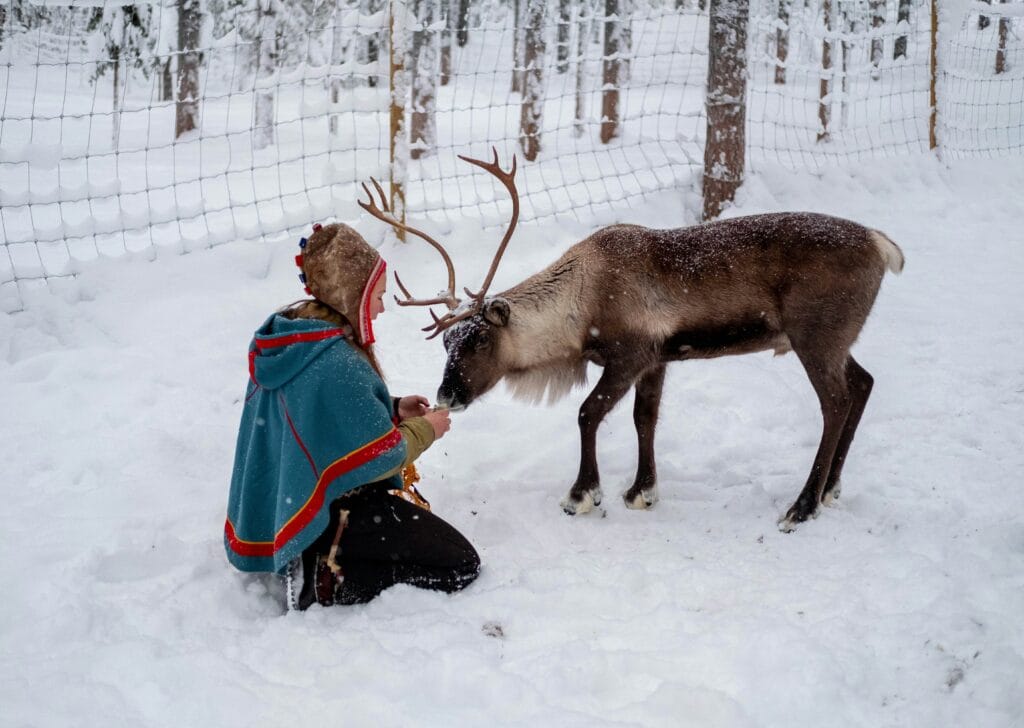 A woman in a traditional blue and red coat kneeling on snow-covered ground to feed a reindeer.