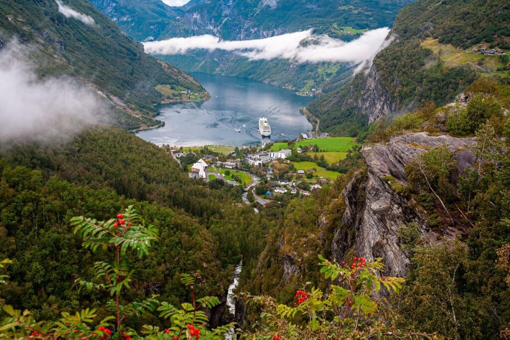 High-angle panoramic view looking down into a deep green Norwegian fjord valley where a white cruise ship sails through mountain cliffs wrapped in low clouds.