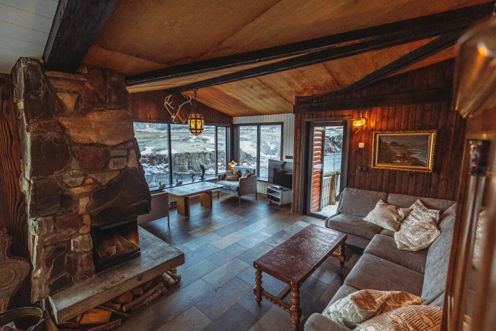 A modern black A-frame cabin glowing with warm golden light from its large glass windows, nestled inside a quiet, snow-covered pine forest at twilight.