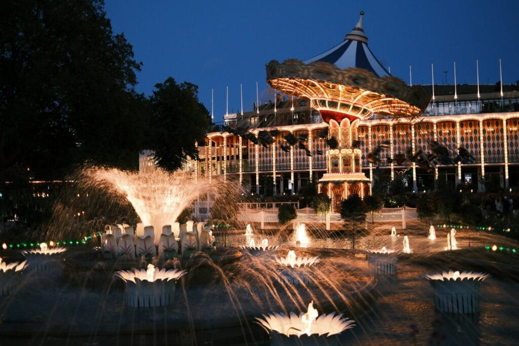 A brightly lit wave swinger carousel spinning at night behind illuminated fountain water displays at Tivoli Gardens theme park in Denmark.