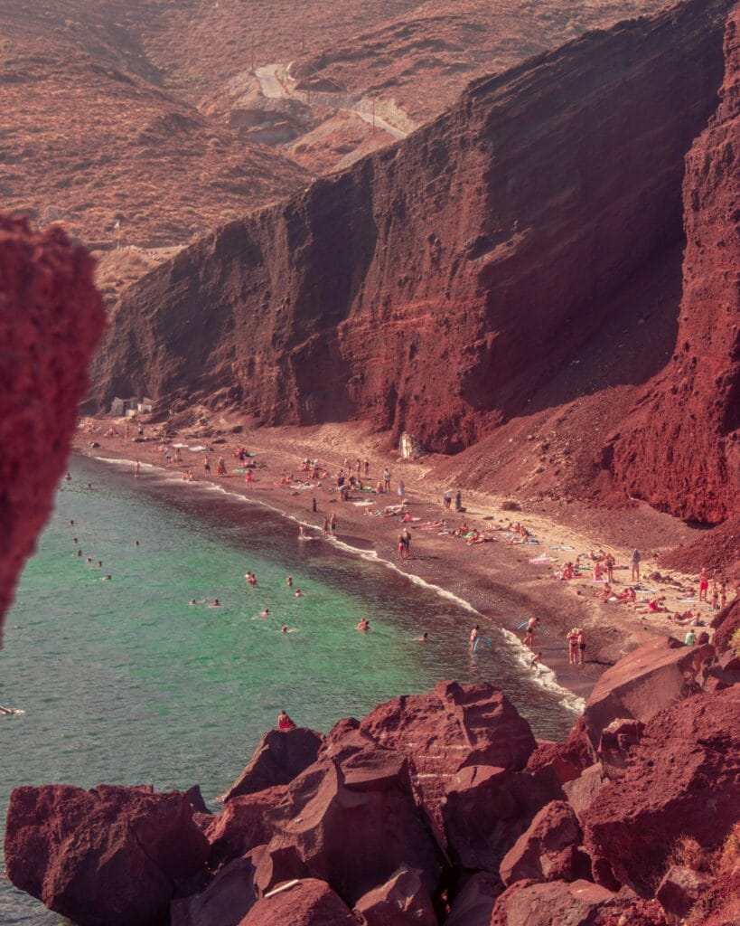 Red sand beach with turquoise water and visitors relaxing along the coastline of Santorini, Greece